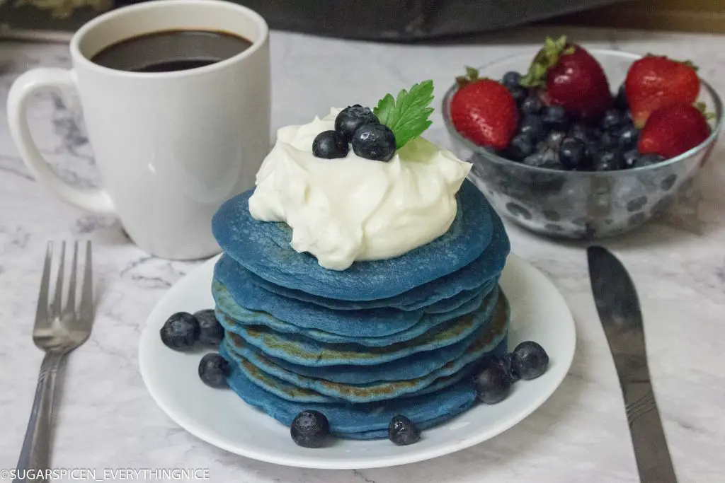 Blue pancakes with whipped cream and blueberries. Coffee and Bowl of strawberries and blueberries in the background
