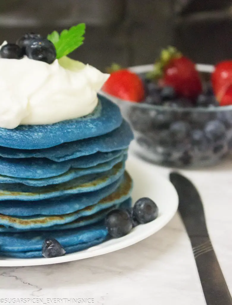 Blue pancakes with whipped cream and blueberries. Coffee and Bowl of strawberries and blueberries in the background
