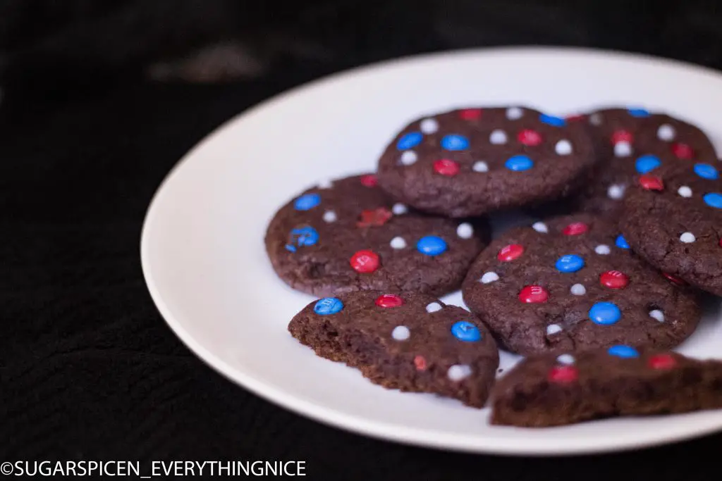 Chocolate cookies with Red, Blue and White MnMs kept on a white plate. On of the cookie is broken into two.