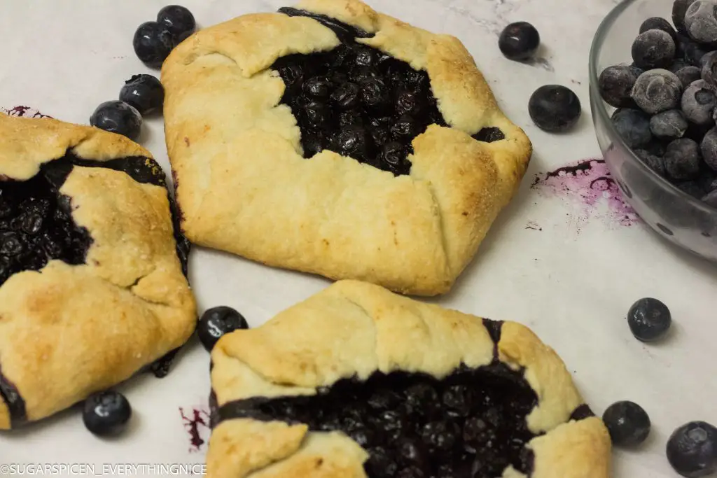 3 blueberry galettes with few blueberry around it placed on white marble countertop.