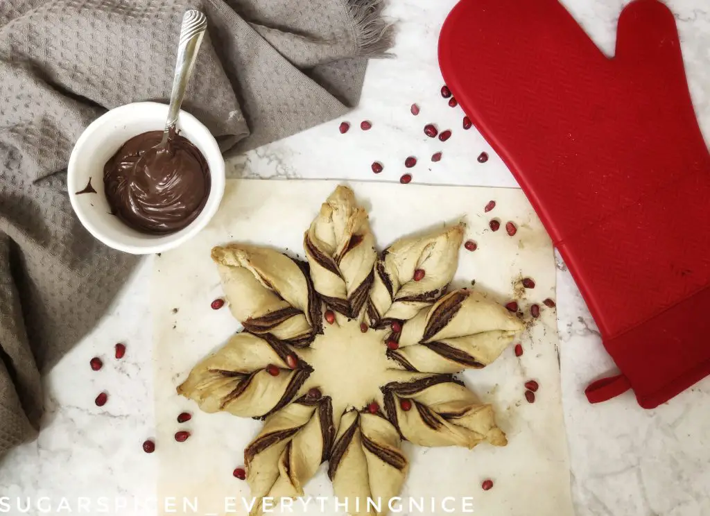 baked star bread with pomegranite seeds over it. A side of vegan chocolate spread and a red oven mitt kept on the side.
