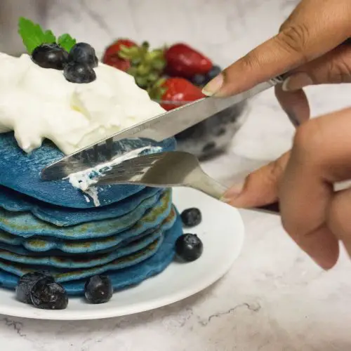 Stack of blue eggless pancakes with whipped cream and blueberries on top, being cut with knife and fork.
