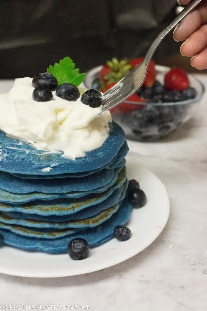 Blue pancakes stacks with whipped cream and blueberries and a fork picking up blueberry. Bowl of strawberries and blueberries in the background.
