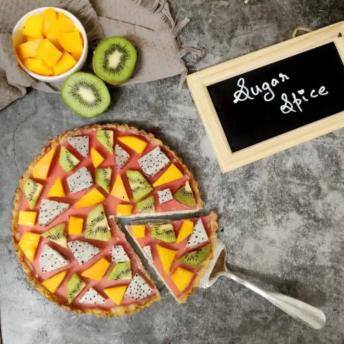 Custard fruit tart with kiwi, dragon fruit and mango arranged in a geometric puzzle pattern over cranberry jelly. A slice is cut from it. Cut mango and kiwi kept on the side as well as a chalkboard with " Sugar Spice" written on it.