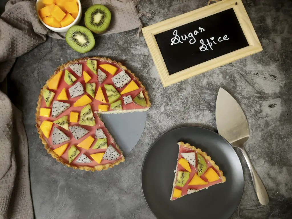 Custard fruit tart with kiwi, dragon fruit and mango arranged in a geometric puzzle pattern over cranberry jelly. A slice is cut from it. Cut mango and kiwi kept on the side as well as a chalkboard with " Sugar Spice" written on it.