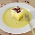 rasmalai cake slice kept on a plate with ras milk around it and a fork kept near it