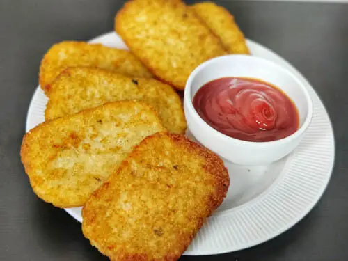 air fryer hash browns in a white plate with a side of ketchup in a small bowl