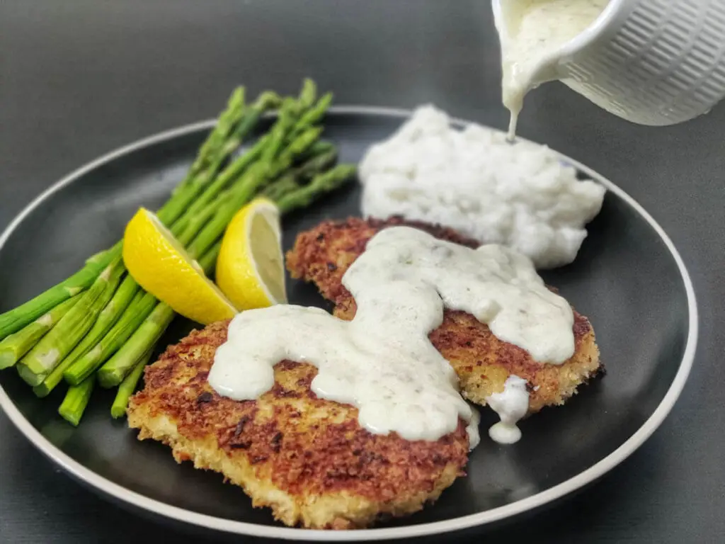 chicken costoletta with lemon sauce poured on it on a black plate with a side of asparagus, mashed potatoes and lemon wedges