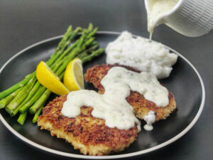 Crispy Chicken Costoletta Cheesecake Factory Recipe chicken costoletta with lemon sauce poured on it on a black plate with a side of asparagus, mashed potatoes and lemon wedges