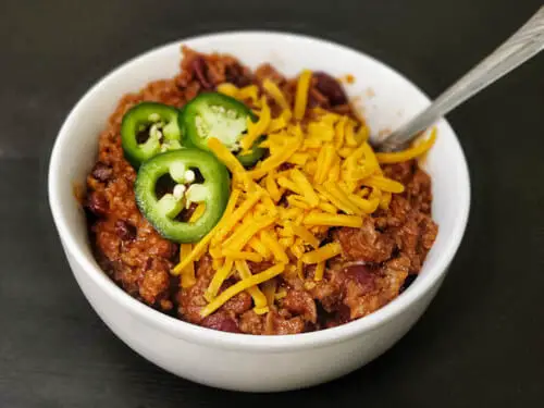 texas roadhouse chili recipe with shredded cheddar and sliced jalapeno in a white bowl and spoon